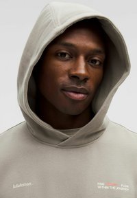 Sweatshirt à capuche gris clair, arborant le logo lululemon et le texte "TROUVEZ LE FLUX DANS LE VOYAGE" en blanc et rouge. Texture douce.