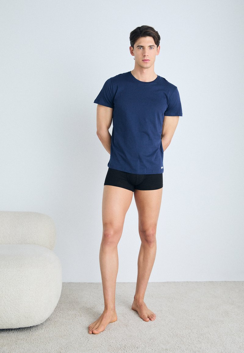 Camiseta de algodón navy combinada con pantalones cortos ajustados negros. Textura suave, mangas cortas, diseño liso. De pie sobre una alfombra clara.