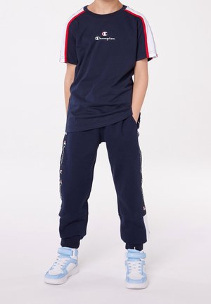 Dziecko ubrane w granatowy dres Champion i białe niebieskie wysokie trampki, stojące z rękami w kieszeniach na białym tle.