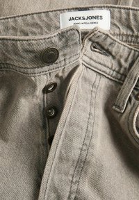 Bežiniai džinsai su mygtukų užsegimu, su tamsiais metaliniais detaliais, matomomis siūlėmis ir audinio etikete, kurioje užrašyta "JACK&JONES."