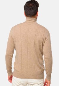 Homme portant un pull à col roulé beige à côtes et un pantalon blanc vu de dos sur un fond blanc uni.