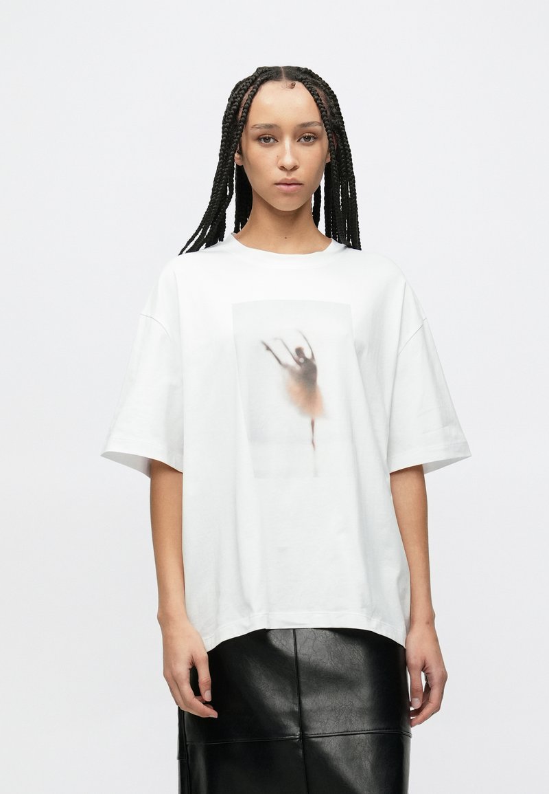 Biała bawełniana koszulka o luźnym kroju, z nadrukiem rozmytego wizerunku tancerza. Okrągły dekolt i krótkie rękawy.
