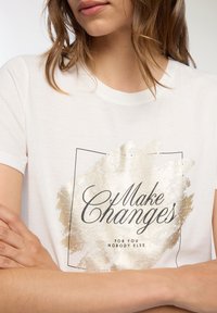 Biała bawełniana koszulka z krótkim rękawem i okrągłym dekoltem, ozdobiona złotym nadrukiem z tekstem "Wprowadź zmiany" oraz prostokątnym konturem.