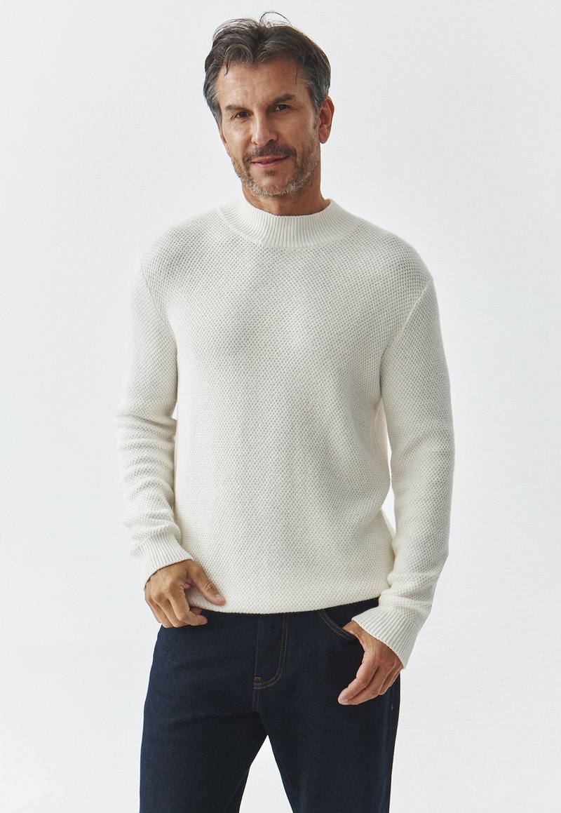 Homme à cheveux courts et foncés, portant un pull en tricot blanc et un jean noir, se tenant avec une main dans la poche devant un fond uni.