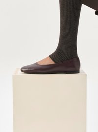 Ballerine in pelle bordeaux con punta arrotondata, suola piatta e un leggero lucido, abbinate a una calza scura a costine su un piedistallo bianco.