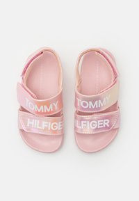 Vaaleanpunaiset sandaalit, joissa on säädettävät hihnat ja valkoinen "TOMMY HILFIGER" -teksti. Pehmeää materiaalia ja teksturoitu pohja; hihnoissa on gradienttiväri-efekti.