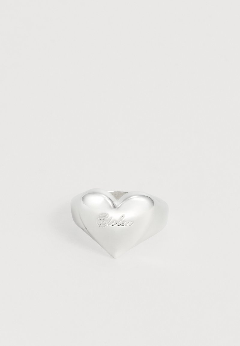 Ασημένιο δαχτυλίδι σε σχήμα καρδιάς με τη λέξη «Stolen» χαραγμένη μπροστά, εμφανίζεται σε απλό ανοιχτό φόντο.