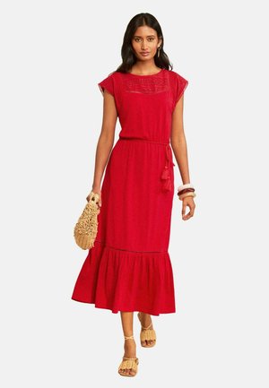 Femme portant une robe rouge sans manches avec une ceinture à nouer, portant un sac tressé et portant des sandales beige et des bracelets.
