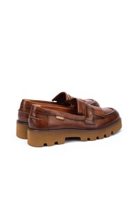 Mocasines de cuero marrón con suela de goma gruesa, que presentan una correa decorativa a lo largo del empeine y un detalle de costura en la parte superior.