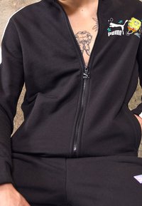 Personne portant une veste zippée noire Puma avec un motif SpongeBob près de l'épaule et un tatouage partiellement visible sur la poitrine.