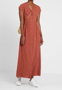 Žena v maxi šatech s knoflíky v rezavé barvě, s krátkými rukávy, bílými teniskami a kulatou crossbody taškou ze slámy.