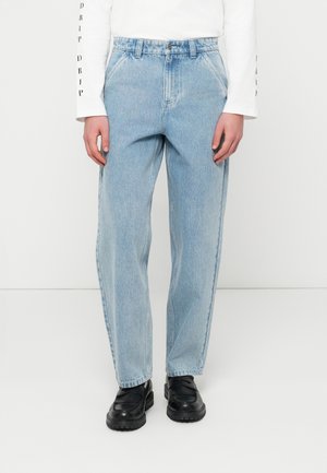 Persona che indossa jeans azzurri a gamba dritta, mocassini neri e una camicia bianca a maniche lunghe con testo sulle maniche, in piedi contro un muro bianco.