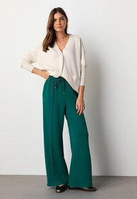 Elfenbeinfarbener Cardigan mit Knöpfen, kombiniert mit grünen weit geschnittenen Hosen mit elastischem Bund. Das Model trägt schwarze Ballerinas. Einfache, lässige Look.