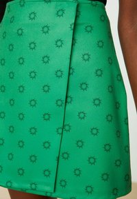 Vihreä hame, jossa on sileä pinta ja toistuva musta aurinkokuosi. Sisältää laskostuksen yksityiskohtana lisäsuunnittelua varten.