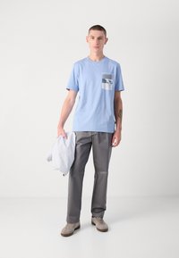 Lyseblå bomull T-skjorte med brystlomme som har et grafisk design, kombinert med grå bukser og lyse sneakers, holder en jakke.