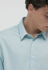 Helles blaues Kurzarmhemd, strukturierter Stoff, Hemdkragen mit Knöpfen, weißen Knöpfen und subtilen Mustern, Nahaufnahme des Kragens und der Schulter.