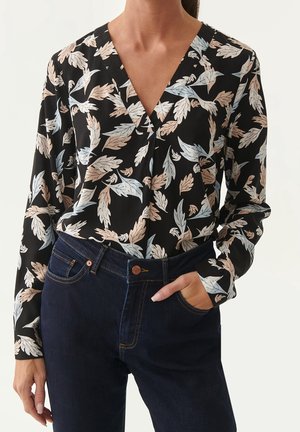 Blouse noire à col en V, avec un motif floral de feuilles bleu clair et beige. Associée à un jean bleu foncé, le modèle a les mains dans les poches.