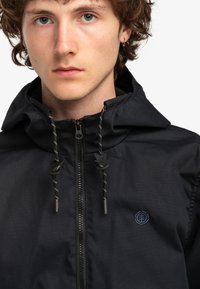 Chaqueta negra con cremallera y capucha, con cordones, un pequeño logo en el pecho y una textura suave y mate.