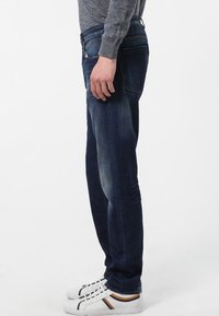 Mörkblå denimjeans med rak passform, med urtvättade detaljer och bakfickor. Kombinerat med vita sneakers och en grå långärmad skjorta.