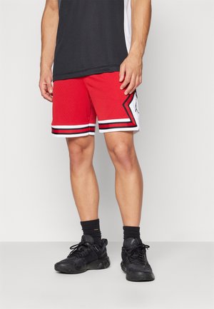 Homme portant un short de basket rouge avec des bordures noires et blanches, des chaussures de sport noires et des chaussettes noires, debout devant un fond clair uni.