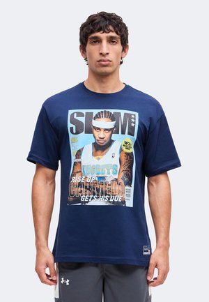 Mann trägt ein dunkelblaues T-Shirt mit einem Basketballmagazin-Cover, das einen Spieler im Nuggets-Trikot und den Text "SLAM" und "Carmelo" zeigt.