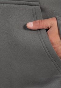 Materiale di felpa grigio con una texture morbida, dotato di tasca frontale e una mano posizionata all'interno della tasca per un dettaglio funzionale.