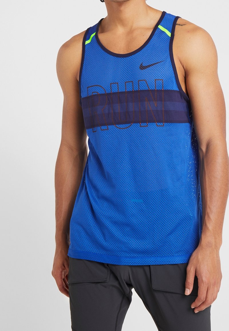 Homme portant un débardeur en mesh bleu Nike avec le texte « RUN » et un short noir, debout devant un fond clair uni.