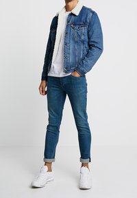Hombre que lleva una chaqueta de denim azul con cuello de sherpa blanco, camiseta blanca, jeans ajustados arremangados y zapatillas blancas, de pie contra un fondo liso.