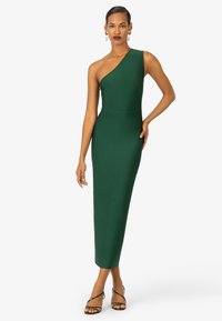 Robe verte, une épaule, ajustée en tissu lisse, midi, avec une silhouette près du corps et des détails de coutures subtils. Sandales à talons fins noires à brides.