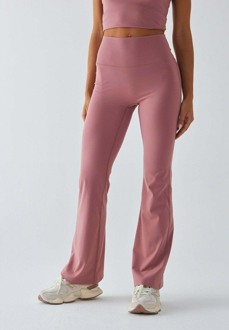 Różowe, rozszerzane legginsy wykonane z gładkiej, elastycznej tkaniny, z wysokim stanem, zestawione z jasnymi butami sportowymi.