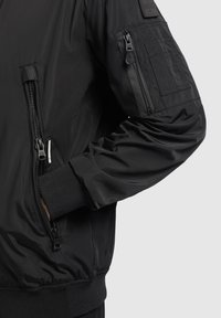 Manche de veste bomber noire avec poignet côtelé, poche zippée sur le bras et main dans la poche latérale zippée sur fond neutre.
