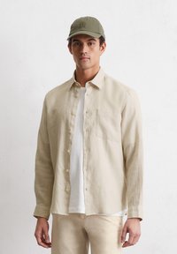 Beige linnen lange mouwen shirt met een knoopsluiting, een enkele zak, en een witte T-shirt met ronde hals eronder; gecombineerd met een groene pet.