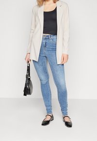 Blazer beige claro, top negro ajustado, jeans skinny azules, bolso negro y zapatos planos negros. Tejidos suaves y diseño ajustado en todo.