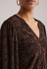 Rjava bluzica z teksturo in V-izrezom, okrašena z rastlinskim vzorcem, dolgimi napihnjenimi rokavi in črnimi gumbi. Gladek material z večplastnim učinkom.