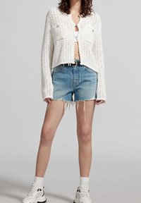 Weißer gestrickter, kurzer Cardigan mit zwei vorderen Taschen, kombiniert mit hellblauen Denim-Shorts. Die Shorts haben ausgefranste Kanten. Weiße Sneakers und Knöchelsocken runden den Look ab.