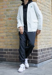 Bílý lehký blejzr přes černou mikinu s kapucí, doplněný černými tepláky a bílými teniskami. V pozadí je texturovaná cihlová zeď.