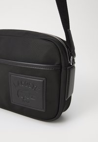Borsa a spalla in tessuto nero con chiusura a zip, dotata di un patch del logo Lacoste testurizzato e di una tracolla larga e decorata. Design rettangolare e liscio.