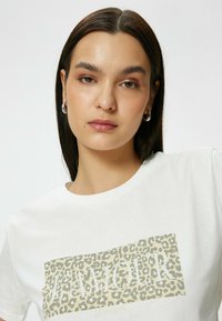 Hvid bomulds T-shirt med et beige leopardprint-design og ordet "AMOUR" i fede bogstaver. Rund halsudskæring og korte ærmer.