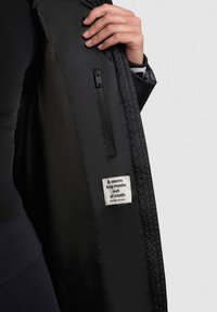 Sort jakke med en glat overflade, der har en lynlås-lomme og et label, der står: "Et varmt kram lavet af stof."