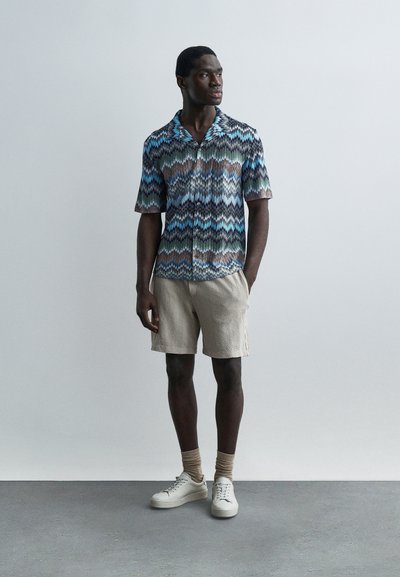 Άνδρας που στέκεται φορώντας μπλε, πράσινο και καφέ πουκάμισο με κοντά μανίκια σε μοτίβο, μπεζ σορτς, μπεζ κάλτσες και λευκά αθλητικά παπούτσια μπροστά σε απλό φόντο.