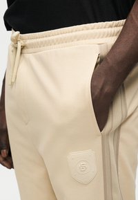 Μπεζ τζόγκερ με λεία υφή, διαθέτουν ελαστική μέση, πλαϊνές τσέπες και ανάγλυφη ετικέτα λογότυπου στον μηρό. Διακοσμημένα με οριζόντιες ρίγες.