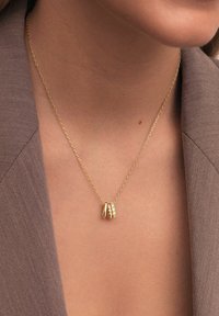 Collier en chaîne doré avec trois petits pendentifs en or, dont un serti de pierres précieuses transparentes, porté sur une peau nue et un blazer marron.