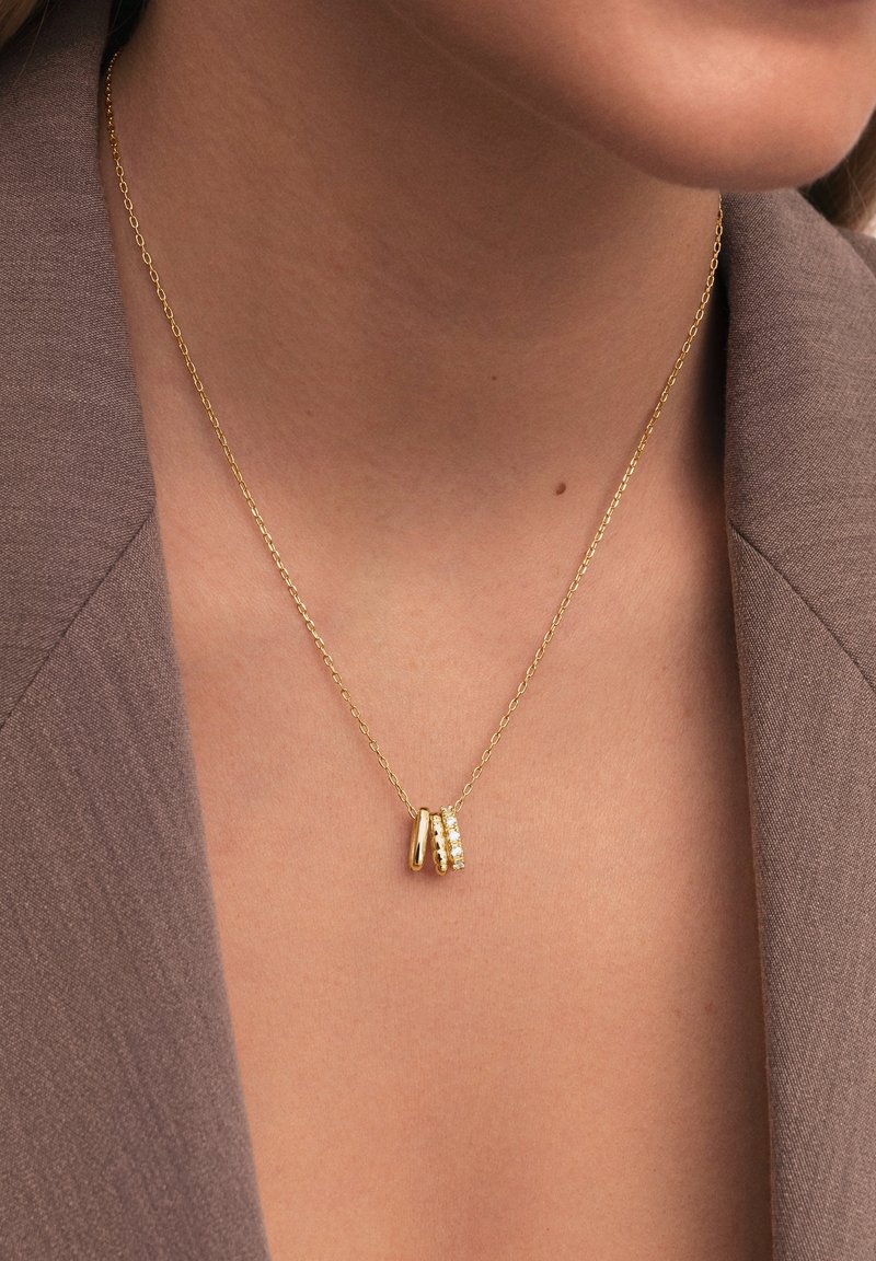 Collier en chaîne doré avec trois petits pendentifs en or, dont un serti de pierres précieuses transparentes, porté sur une peau nue et un blazer marron.