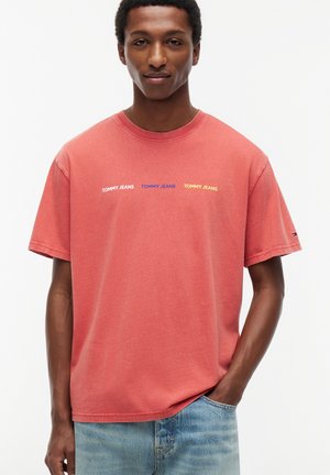 Homme portant un t-shirt rouge délavé avec "TOMMY JEANS" imprimé trois fois en blanc, bleu et jaune, associé à un jean bleu clair.