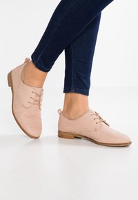 Chaussures en cuir rose clair avec des lacets, texture douce, petit talon et bout arrondi. Associées à un jean ajusté bleu foncé.