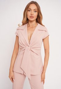 Woman with long blonde hair wearing light pink short-sleeve blouse with front knot and matching pants, standing against a plain light background.