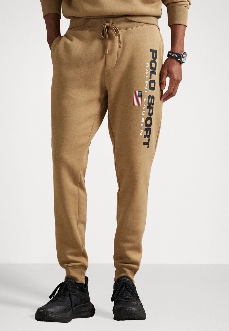 Pantaloni de trening bej cu talie elastică, șnur și buzunare laterale. Caracteristici grafica "POLO SPORT" și detalii cu steagul american pe piciorul stâng.