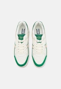 Białe sneakersy z zielonymi akcentami i beżowymi detalami, z perforowanym przodem, płaskimi sznurowadłami oraz logo na języku. Miękka tekstura.