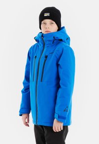 Blaue wasserdichte Jacke mit Kapuze, schwarzen Reißverschlusstaschen und verstellbaren Bündchen. Schwarze Mütze mit Logo. Glatte Textur, für Wärme konzipiert.