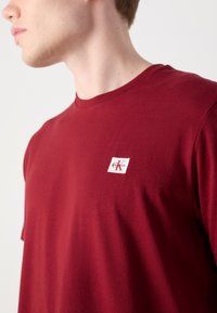 Bordó T-shirt, puha pamutból készült, kerek nyakkivágással és bal mellen egy kis márkázott címkével, fehér háttérrel és piros logóval.